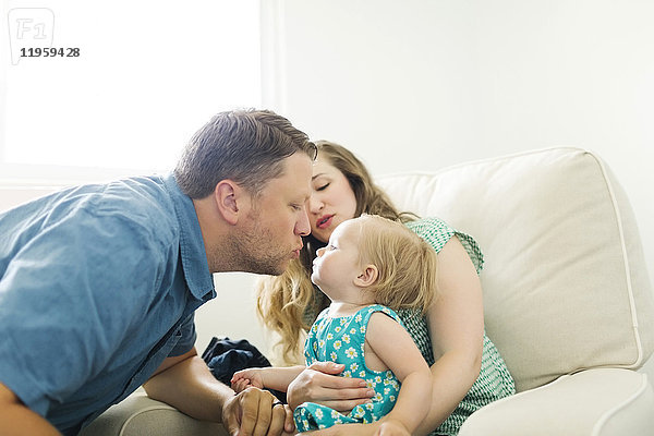 Mutter und Vater mit einem kleinen Mädchen (12-17 Monate) sitzen im Wohnzimmer und küssen sich