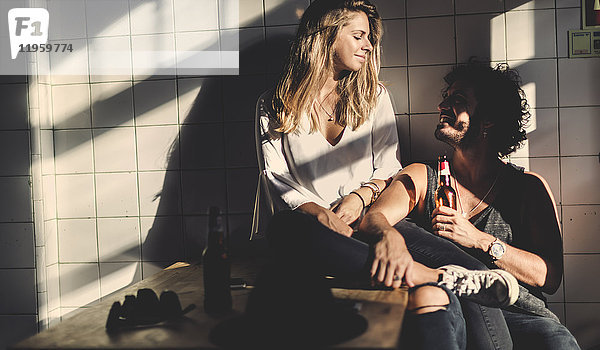 Mann mit braunen Haaren  der eine Bierflasche hält  und eine Frau mit langen blonden Haaren sitzen drinnen  schauen sich an und lächeln.