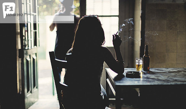 Rückansicht der Silhouette einer Frau  die drinnen an einem Tisch sitzt und Zigarette  Bierglas und Flasche auf dem Tisch raucht.