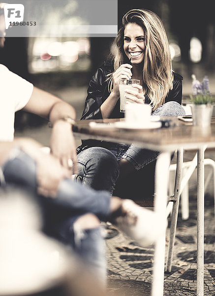 Mann und Frau mit langen blonden Haaren sitzen draußen an einem Tisch in einem Café  die Frau hält ein Getränk in der Hand und lächelt einander an.