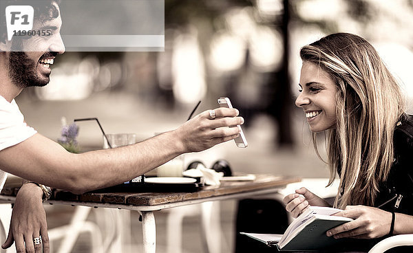 Bärtiger Mann mit Sonnenbrille und Frau mit langen blonden Haaren sitzt draußen an einem Tisch in einem Cafe  Frau schaut auf Smartphone  das der Mann in der Hand hält.
