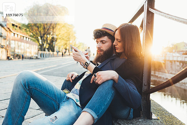 Hipster-Paar sitzt am Stadtkanal und sieht sich ein Smartphone an