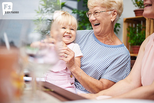 Ältere Frau lacht mit Kleinkind-Enkelin beim Familienessen auf der Terrasse