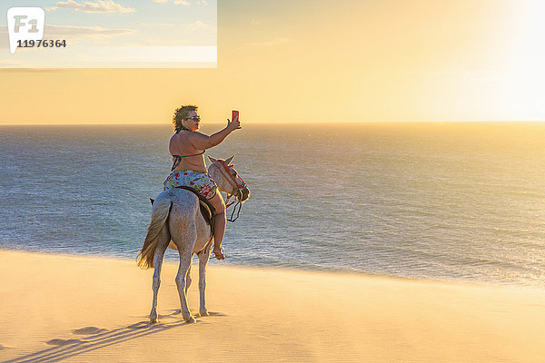 Frau reitet auf einem Pferd am Strand  fotografiert die Aussicht mit einem Smartphone  Jericoacoara  Ceara  Brasilien  Südamerika