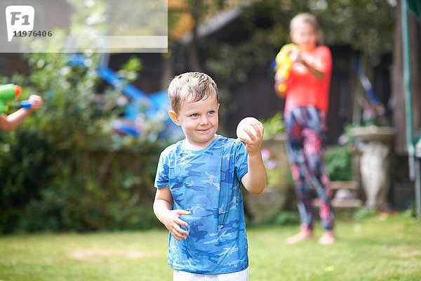 Spitzbübischer Junge hält Wasserballon im Garten