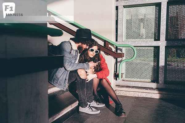 Junges Paar sitzt auf einer Treppe und schaut auf ein Smartphone