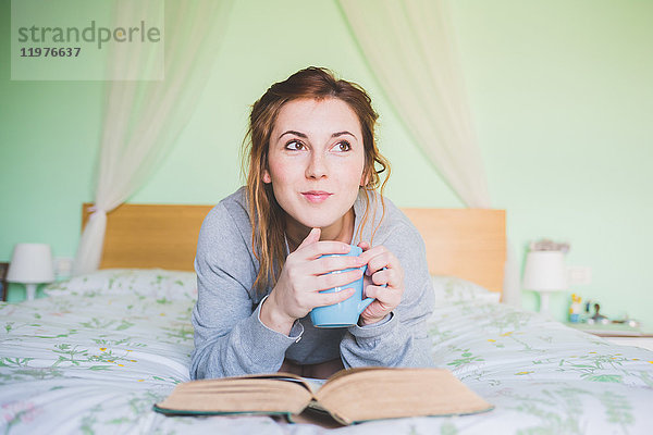 Junge Frau liegt auf dem Bett  hält Kaffeetasse und schaut auf