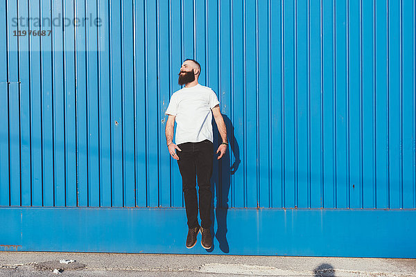 Bärtiger Mann vor blauer Wand springt in die Luft