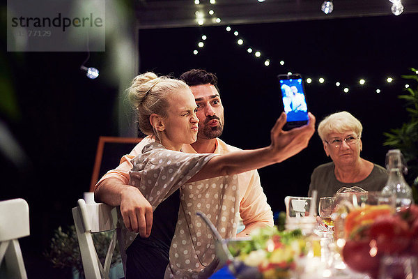 Gruppe von Menschen sitzt am Tisch  genießt die Mahlzeit  junge Frau nimmt sich selbst mit dem Smartphone