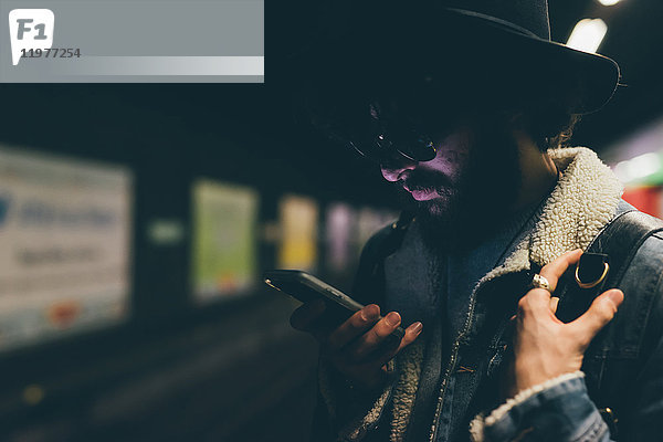Junger Mann steht auf U-Bahn-Bahnsteig und schaut auf Smartphone