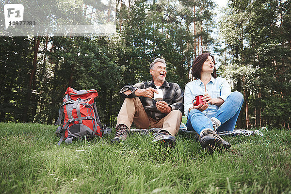 Ein erwachsenes Paar entspannt sich im Gras  hält Blechbecher  Rucksack daneben