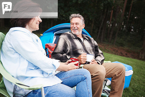 Ein erwachsenes Paar sitzt auf Campingstühlen  neben dem Zelt  hält Tassen Tee und lächelt