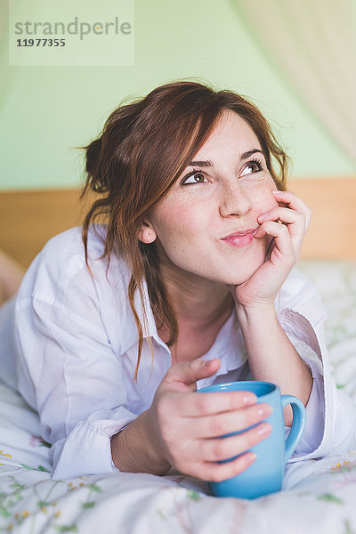Junge Frau im Bett mit Kaffeetasse  nach oben schauend