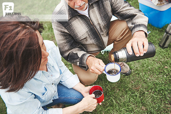 Reifes Paar im Gras kauernd  Mann gießt heißes Getränk aus Getränkeflasche aus  erhöhte Ansicht