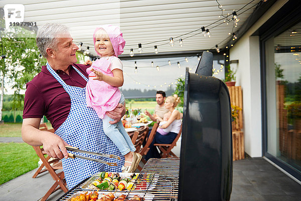 Erwachsener Mann trägt Kleinkind-Enkelin beim Familienessen auf der Terrasse grillen