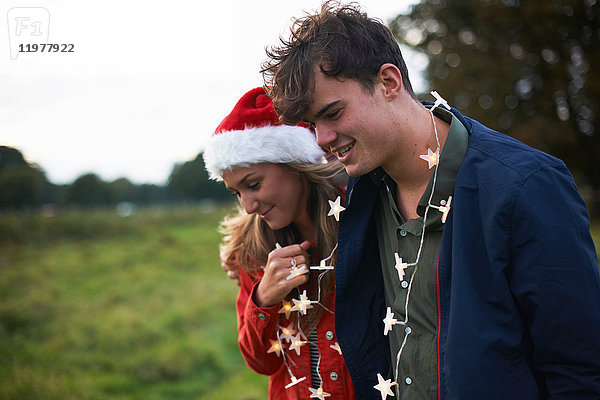 Junges Paar mit Weihnachtsmannhut und Weihnachtslichtgirlande auf dem Feld spazieren