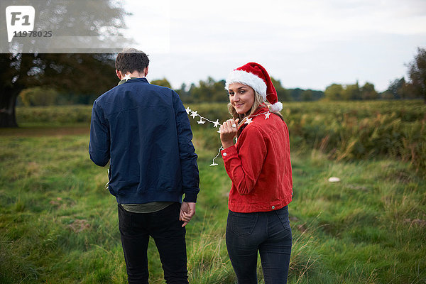 Rückansicht eines jungen Paares mit Weihnachtsmannhut und Weihnachtslichtgirlande beim Spaziergang im Feld
