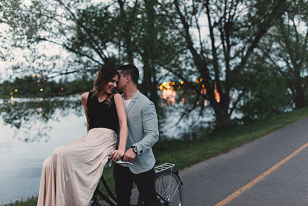 Romantischer junger Mann flüstert in der Dämmerung am See am Fahrradlenker seiner Freundin zu
