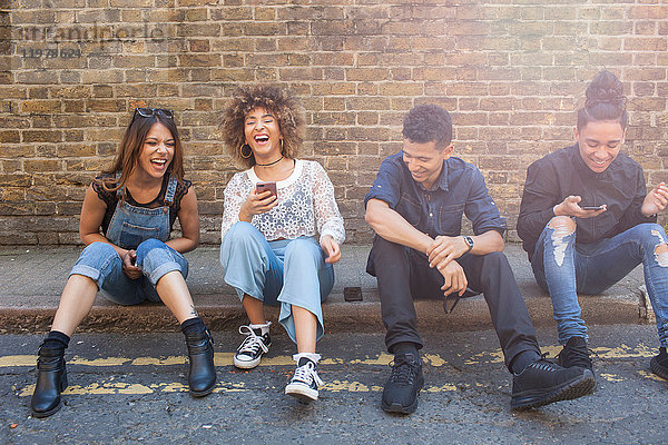 Vier Freunde sitzen auf der Straße  lachend  junge Frau mit Smartphone