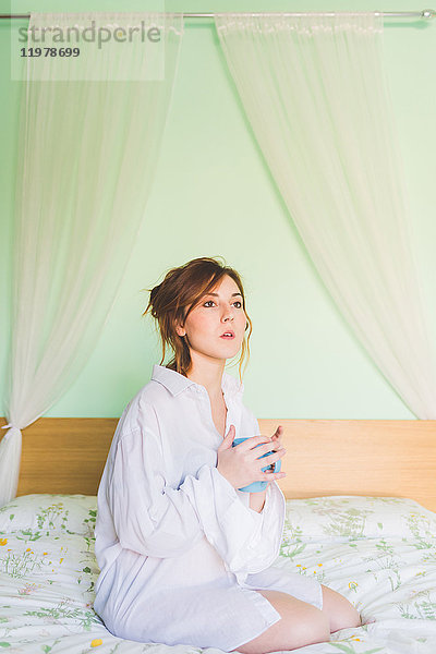 Junge Frau kniet auf Bett  hält Kaffeetasse und starrt