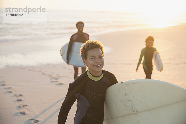 Portrait lächelnder vorpubertärer Junge im Neoprenanzug mit Surfbrett am Sommer-Sonnenuntergangstrand mit Familie