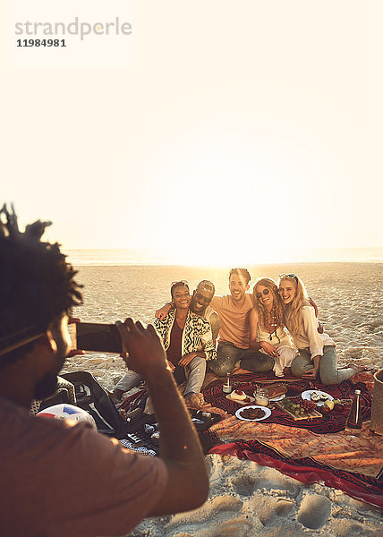 Junger Mann mit Fotohandy fotografiert Freunde beim Picknick am sonnigen Sommerstrand