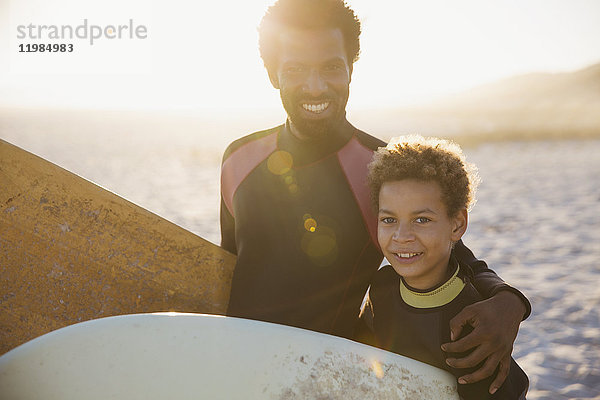 Portrait lächelnder  selbstbewusster Vater und Sohn in Neoprenanzügen mit Surfbrettern am sonnigen Sommerstrand.