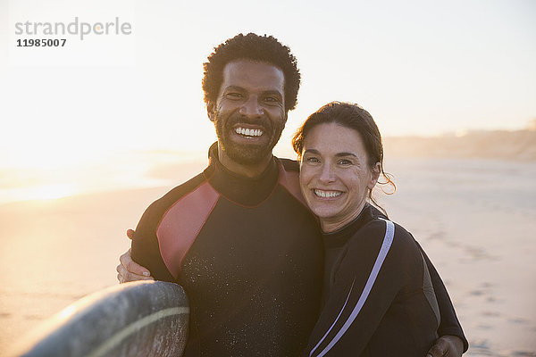 Portrait lächelndes  selbstbewusstes multiethnisches Paar mit Surfbrett am sonnigen Sommersonnenuntergangstrand.