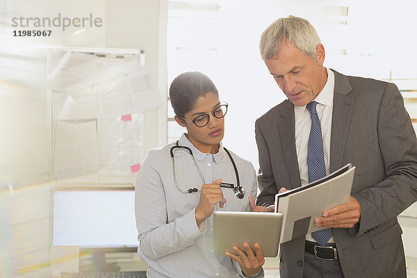 Ein weiblicher Arzt und ein männlicher Krankenhausverwalter unterhalten sich  schauen auf ein digitales Tablet und Papierkram im Untersuchungsraum