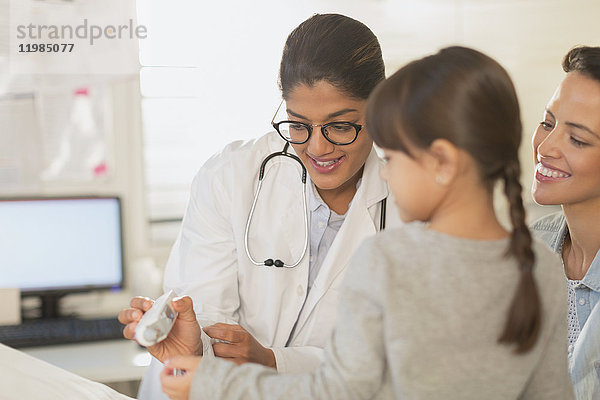 Eine Kinderärztin zeigt einem Mädchen und seiner Mutter im Untersuchungsraum ein digitales Thermometer