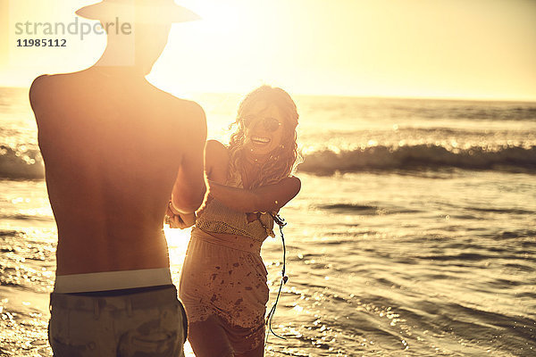 Verspieltes junges Paar hält sich am sonnigen Sommer-Sonnenuntergang am Meer fest