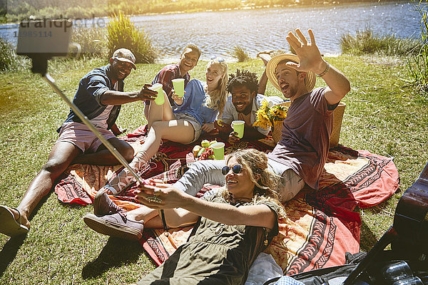 Verspielte junge Freunde nehmen Selfie mit Selfie-Stick  genießen Picknick am sonnigen Sommerufer