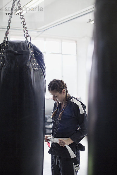 Junge Frau beim Binden des Judogürtels neben dem Boxsack im Fitnessstudio