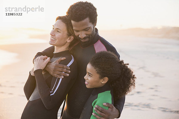 Liebevolle multiethnische Familie in Neoprenanzügen am Strand bei Sonnenuntergang
