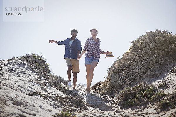 Verspieltes  energiegeladenes  multiethnisches Paar  das auf einem sonnigen Sommerstrandweg läuft und Händchen hält.