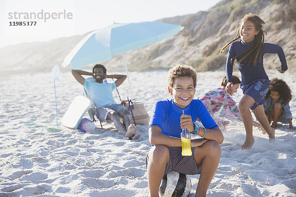 Portrait lächelnder vorpubertärer Junge trinkt Saft am sonnigen Sommerstrand mit Familie