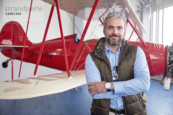 Portrait lächelnder  selbstbewusster männlicher Flugzeugpilot am Doppeldecker im Hangar stehend