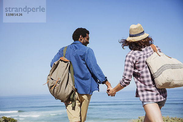 Zärtliches multiethnisches Paar  das sich an den Händen hält und am sonnigen Sommerstrand spazieren geht.