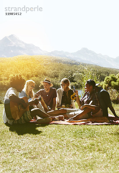 Junge Freunde hängen herum  spielen Gitarre und genießen ein Picknick im sonnigen Sommergras.