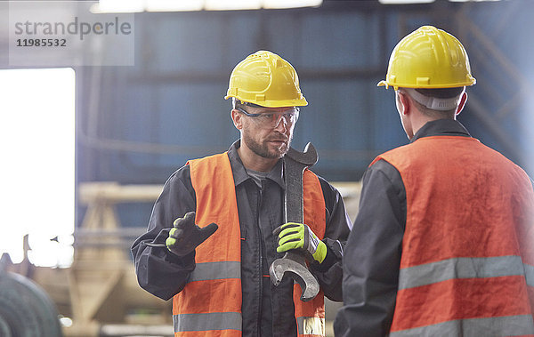 Arbeiter mit großem Schraubenschlüssel im Gespräch mit Kollegen in der Fabrik