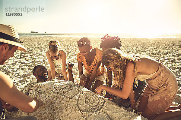 Junge Freunde begraben und zeichnen Freund im Sand am sonnigen Sommerstrand