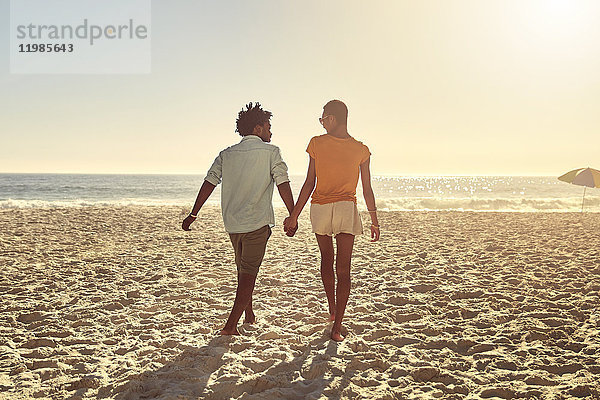 Junges Paar hält sich an den Händen und geht am sonnigen Sommerstrand spazieren.