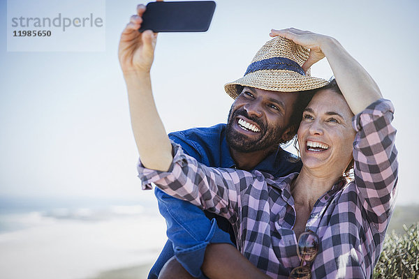 Verspieltes  lächelndes multiethnisches Paar  das sich selbst mit Fotohandy am sonnigen Sommerstrand fotografiert.