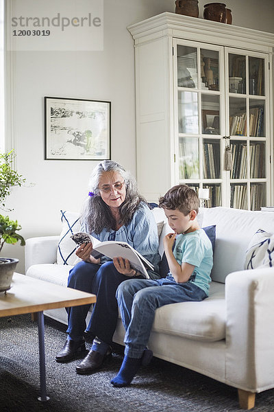 Großmutter und Enkel lesen Bilderbuch sitzend auf dem Sofa zu Hause