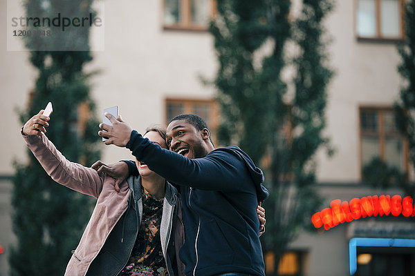 Fröhliche junge Freunde  die Selfie von Smartphones nehmen  während sie gegen das Gebäude stehen