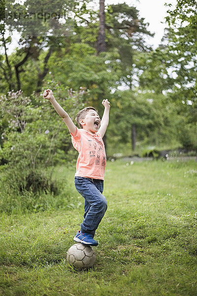 Aufgeregter Junge schreit mit erhobenen Armen  während er auf einem Fußball im Hinterhof steht.