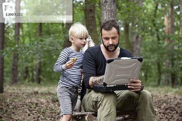 Vater und Sohn sitzen auf einem selbstgebauten Holzstuhl im Wald mit Laptop