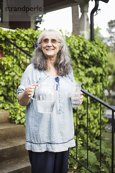 Lächelnde Seniorin stehend mit Wasserkrug und Glas im Garten