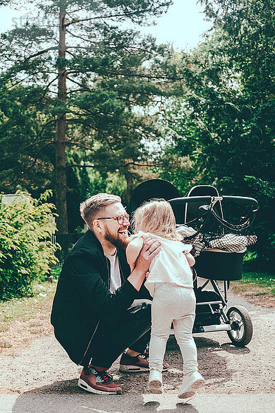 Glücklicher Mann hockt  während er seine Tochter mit dem Kinderwagen auf dem Fußweg im Park hält.