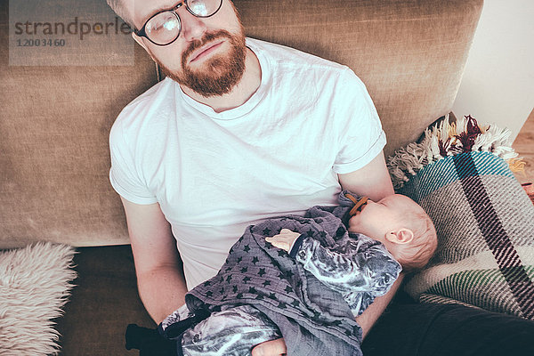 Hochwinkelansicht des Mannes  der schläft  während er das Baby auf dem Sofa hält.
